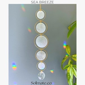 May include: A hanging ornament with a gold chain and circular frames. Each frame holds a white, iridescent disc. A clear crystal hangs at the bottom. The text "SEA BREEZE" and "Solmate.co" are visible. The ornament is for decoration.