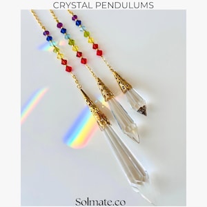 May include: Three crystal pendulums with gold-colored filigree caps and chains. Each pendulum has a clear, faceted crystal point and a strand of colorful beads. The image includes the text "CRYSTAL PENDULUMS" and the website "Solmate.co".