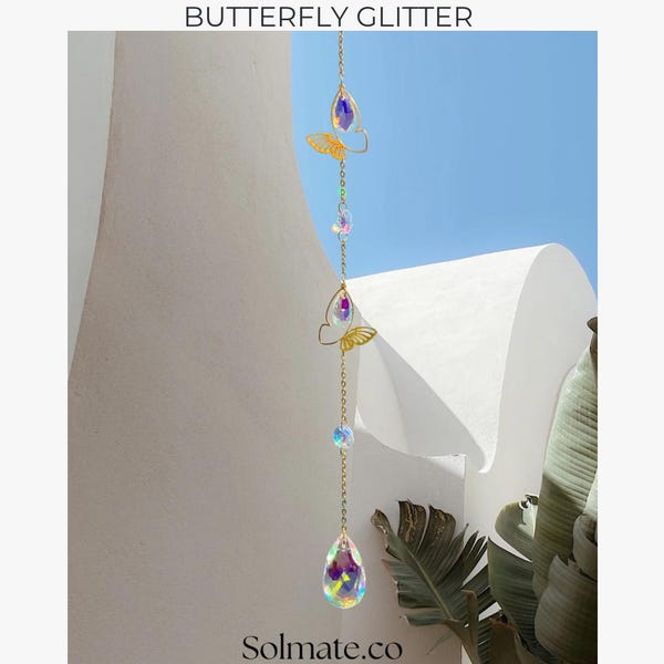 May include: A hanging ornament with the text "BUTTERFLY GLITTER" at the top. The ornament features gold-colored butterfly-shaped accents and iridescent crystal teardrops and beads suspended on a delicate chain. The background includes white architectural elements and a blue sky.