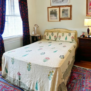 May include: A bedroom with a white bedspread with floral patterns, a wooden nightstand with a lamp, and a rug with a blue, red, and white pattern.