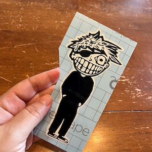 May include: Black and white sticker of a cartoon character with spiky hair, wearing a black shirt and trousers, and a single eye with a mischievous grin. The sticker is on a blue gridded transfer sheet.