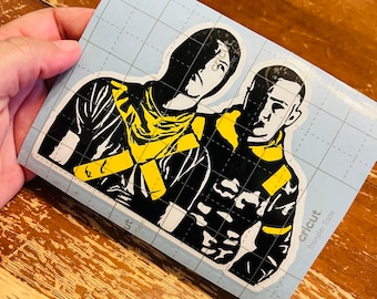 Può includere: Decalcomania in vinile nero e giallo di due figure con le braccia intorno l'una all'altra. La decalcomania è su un foglio di trasferimento a griglia blu con il logo Cricut e le parole "Transfer Tape".