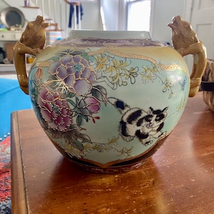 Puede incluir: Un jarrón de porcelana con detalles dorados y diseños florales. El jarrón tiene dos asas con forma de rana y presenta un diseño de gato blanco y negro.