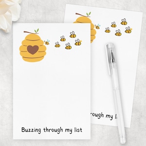 May include: Two white notepads with a bee-themed design. Each notepad features a yellow beehive with a heart, cartoon bees, and the text "Buzzing through my list." A clear pen rests on one notepad.