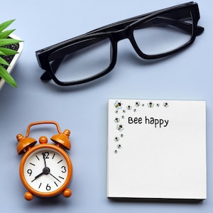 May include: An orange alarm clock, black-framed glasses, and a small potted plant sit on a light blue surface. A white notepad with the words "Bee happy" and bee illustrations is also present. The clock shows the time.