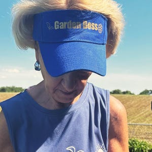 May include: A blue visor with the text "Garden Boss" in gold lettering. The person is wearing a blue tank top with a turtle and sun graphic. The background includes a field and blue sky.