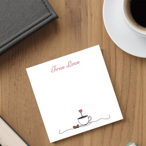 May include: A white notepad with the words "True Love" in pink script. A minimalist line drawing of a coffee cup with a heart above it and coffee beans is below the text. A cup of coffee sits on a white saucer.