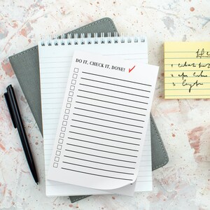 May include: A top-down view of a desk setup featuring a black pen, a gray notebook, and a white spiral-bound notepad with the text "DO IT, CHECK IT, DONE!" and a red checkmark. A yellow sticky note with handwritten notes is also visible.