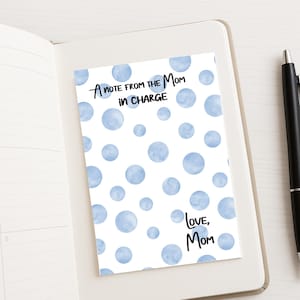 May include: A white card with blue watercolor polka dots and the text "A note from the Mom in Charge" and "Love, Mom". The card is on a white notebook with a black pen.