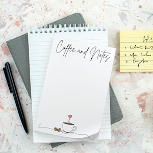May include: A white notepad with the words "Coffee and Notes" in script, a coffee cup illustration, and a small red heart. A black pen and a yellow sticky note with handwritten notes are also visible.