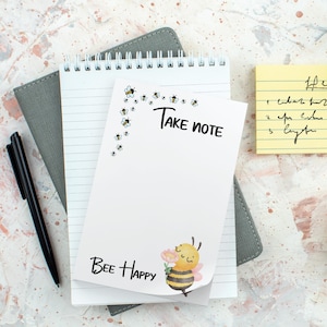 May include: A white spiral notebook with the text "Take Note" and "Bee Happy" printed on the cover. The cover features a cartoon bee holding a flower and small bee illustrations. A black pen and a yellow sticky note are also visible.