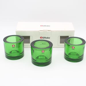 Op de afbeelding: Drie cilindrische votiefkaarsenhouders van groen glas met dikke wanden en een ronde opening. Elke houder is voorzien van een kleine rood-witte iittala-logosticker. Achter de houders staat een witte kartonnen doos met het iittala-logo en de tekst KIVI.