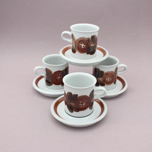 Könnte beinhalten: Ein Set aus vier weißen Keramiktassen und Untertassen, die gestapelt sind. Jede Tasse und Untertasse zeigt ein braunes Blumenmuster mit kreisförmigen Zentren und dunkelbraunen Blättern. Die Untertassen haben einen soliden braunen Rand.
