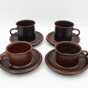 Peut inclure: Quatre tasses à café et soucoupes en céramique marron. Les tasses sont cylindriques avec de petites anses. Les soucoupes sont rondes avec un bord légèrement surélevé. L'ensemble a une esthétique rustique et terreuse.