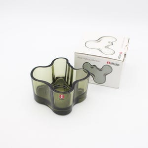 Op de afbeelding: Een mosgroene glazen kom met een golvende, organische, meerlobbige rand en een gladde basis. De kom is voorzien van een kleine rood-witte Iittala merksticker. Daarnaast staat een witte kartonnen doos met het opschrift Alvar Aalto Collection en Iittala.