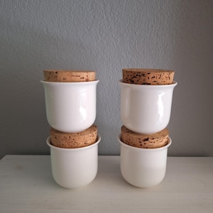 Puede incluir: Cuatro tarros de cerámica blancos con tapas de corcho apilados en dos pares. Los tarros tienen un acabado brillante y una forma redondeada. Las tapas de corcho son de color marrón claro y encajan perfectamente en los tarros. Los tarros están sobre una superficie blanca.