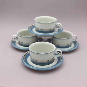 Op de afbeelding: Een set van vier off-white keramische kopjes en schoteltjes. Elk stuk is voorzien van horizontale banden in blauwe en bruine tinten. De kopjes hebben een afgerond oortje en een gladde afwerking, terwijl de schoteltjes een bijpassend cirkelvormig patroon hebben.