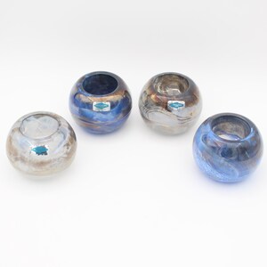 May include: Four spherical glass votive holders with swirling patterns in shades of blue, brown, and white. Each piece features a smooth, rounded opening at the top and a small, rectangular teal sticker with white text attached to the exterior surface.