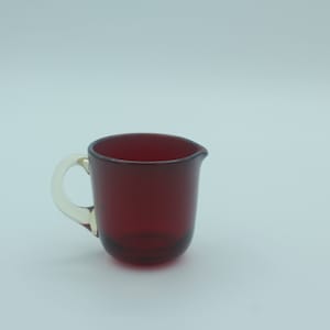 May include: A small, ruby-red glass creamer with a clear handle. The creamer has a rounded base and a spout for easy pouring. The handle is a contrasting clear glass. Suitable for serving cream or milk.