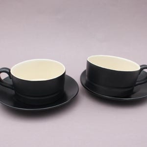 Puede incluir: Dos tazas de cerámica con sus platos a juego, cada una con un exterior negro mate y un interior liso de color crema. Las tazas tienen una forma redondeada con un asa lateral y descansan sobre platos circulares con el mismo acabado negro.