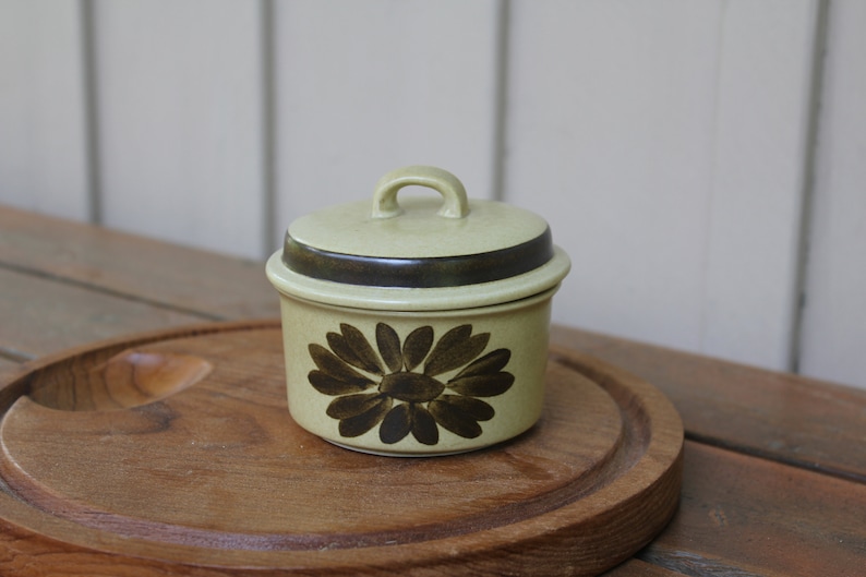 Pode incluir: Um pequeno a&ccedil;ucareiro redondo de cer&acirc;mica com tampa. O a&ccedil;ucareiro &eacute; decorado com um desenho floral castanho e verde. A tampa tem uma faixa castanha em volta da parte superior.