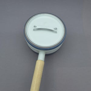 Puede incluir: Una cacerola blanca con tapa y mango de madera. La cacerola tiene un borde azul y un mango blanco. La tapa tiene un pequeño asa. La cacerola está sobre una superficie gris.