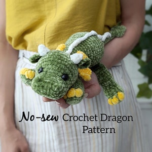 Könnte beinhalten: Ein grünes gehäkeltes Drachenspielzeug mit weißen Hörnern und gelben Akzenten. Der Drache wird in den Armen gehalten. Der Text "No-sew Crochet Dragon Pattern" befindet sich am unteren Rand des Bildes.
