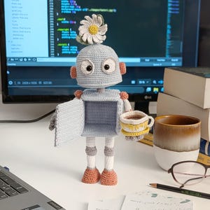 May include: A crocheted robot holding a small crocheted cup. The robot is light blue with a white chest and has a daisy on its head. The robot is standing on a white surface with a laptop and books in the background.