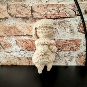May include: A crocheted doll with a cream-colored body and a white crocheted hat. The doll is standing on a black surface.
