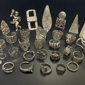 May include: A collection of silver-toned rings with various designs, including geometric shapes, floral patterns, and abstract motifs. The rings are arranged on a dark background.