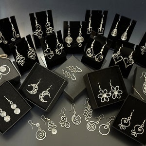 May include: Assortment of silver wire earrings displayed on black velvet boxes. The earrings feature various designs, including spirals, hearts, moons, and flowers.