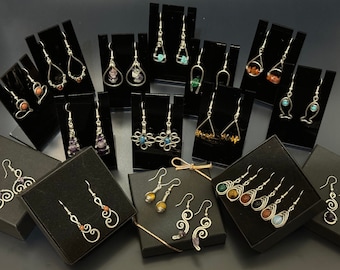 May include: Assortment of handmade earrings with various gemstone and wire designs displayed on black stands. The earrings feature a variety of colors, including turquoise, purple, brown, and yellow.