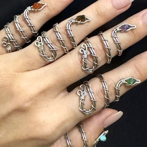 May include: A set of seven silver snake rings with different colored gemstones. The rings are wrapped around the fingers of a hand.