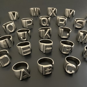May include: A set of 26 silver rings, each featuring a different letter of the alphabet. The rings are arranged in a circle on a dark gray surface.