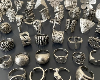 May include: A collection of silver-toned rings with various designs, including geometric shapes, animal motifs, and abstract patterns. The rings are arranged on a dark gray surface.