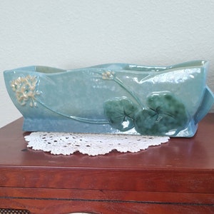 May include: A rectangular, pale blue ceramic planter with a glossy finish. The planter has embossed floral and leaf designs in green and cream. It rests on a vintage wooden stereo, with a white doily beneath it.