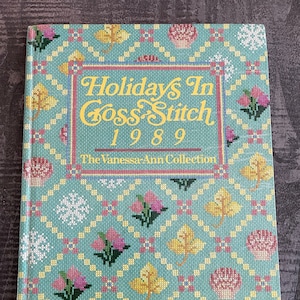 Peut inclure: Un livre bleu turquoise avec un motif de point de croix sur la couverture. Le titre est "Holidays In Cross Stitch 1989" et le sous-titre est "The Vanessa Ann Collection".