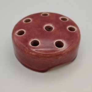 May include: A small, round, red ceramic flower vase with seven holes for stems. The vase has a slightly uneven base.