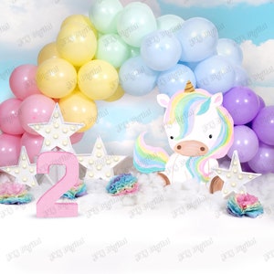 May include: A pink number two with white stars and a unicorn with a rainbow mane and tail. The unicorn is sitting on a white fluffy surface with pastel balloons in the background.