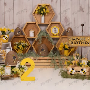 Puede incluir: Un conjunto de decoración de fiesta de cumpleaños con temática de abejas con un número dos amarillo, una caja de madera con un diseño de abeja y un letrero que dice "Hap-Bee 2nd Birthday". Las decoraciones están dispuestas sobre un fondo de madera con estantes de panal y acentos florales.