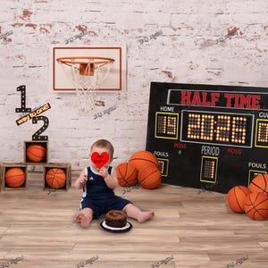 May include: A basketball-themed photo setup with a miniature hoop, scoreboard, and basketballs. A baby in a navy blue outfit sits near a small cake. The scoreboard reads "HALF TIME" and displays scores. Wooden blocks spell out "1/2 way to one!"