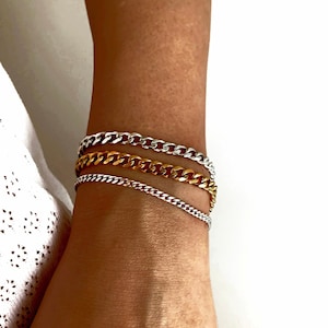 May include: A set of three chain bracelets in silver and gold tones. The bracelets are layered on a person's wrist.