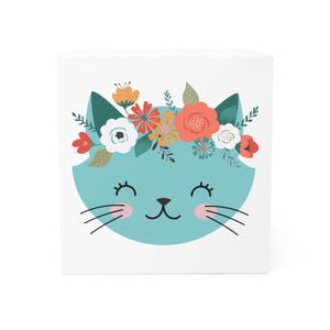 May include: A white square block with a cartoon cat face in light blue, adorned with a floral crown. The cat has closed eyes, pink cheeks, and black whiskers. The crown features orange, red, and white flowers.