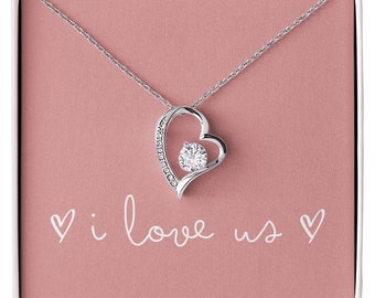 May include: A silver necklace with a heart-shaped pendant featuring a sparkling clear stone. The pendant is set in a delicate open heart design with smaller stones around the edge. The necklace comes in a gift box with the words "i love us" printed on the front.