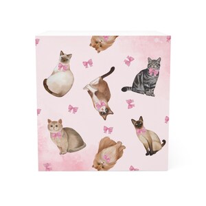 May include: A decorative cube with a repeating pattern of watercolour cats in different poses. Each cat wears a pink bow. The background is a soft pink with scattered pink bow accents.