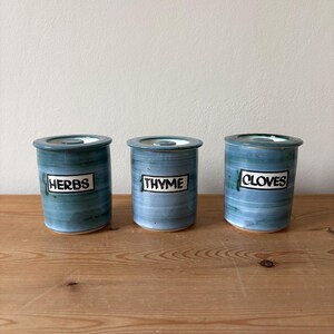 May include: Three ceramic spice jars with lids, labelled "HERBS", "THYME", and "CLOVES". The jars are cylindrical with a blue and green marbled glaze. They sit on a wooden surface.