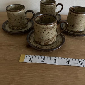May include: Set of four brown and beige speckled ceramic coffee cups with matching saucers. The cups have a rounded shape and a handle on the right side. A measuring tape is in the foreground.