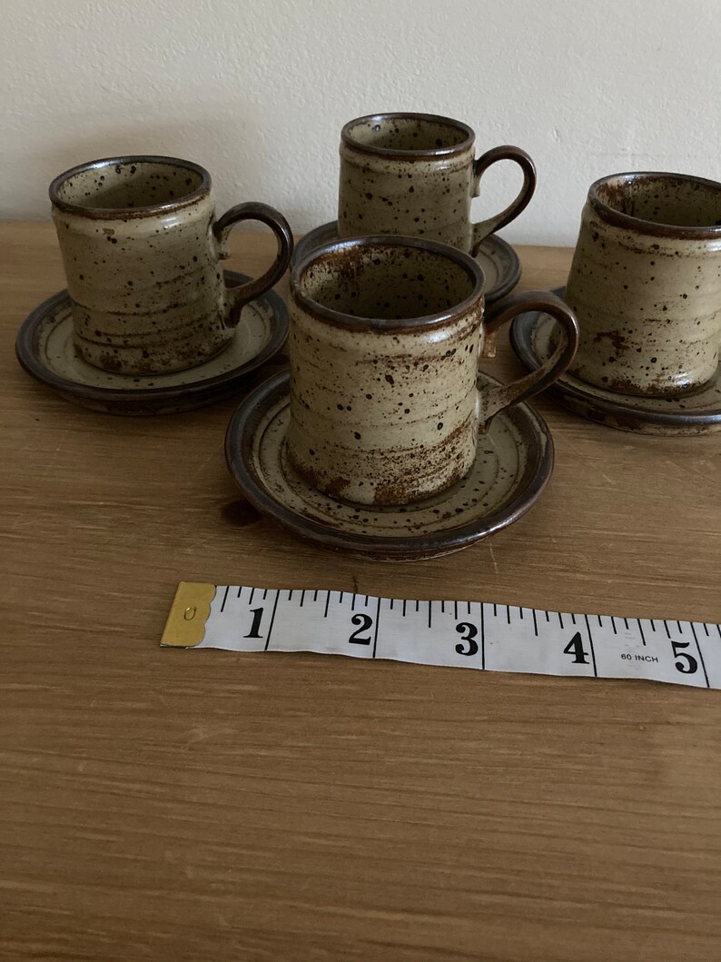May include: Set of four brown and beige speckled ceramic coffee cups with matching saucers. The cups have a rounded shape and a handle on the right side. A measuring tape is in the foreground.