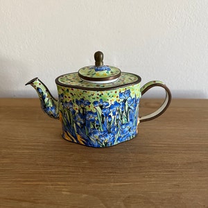 May include: A small green teapot with a floral design featuring blue flowers and yellow accents. The teapot has a brass lid and a brass spout.