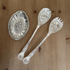 May include: Two silver serving spoons with ornate floral designs and a matching silver serving dish.